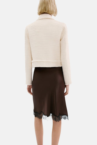 Een vrouw van achteren gezien, gekleed in een crèmekleurig jasje en een bruine rok met zwarte kanten rand.