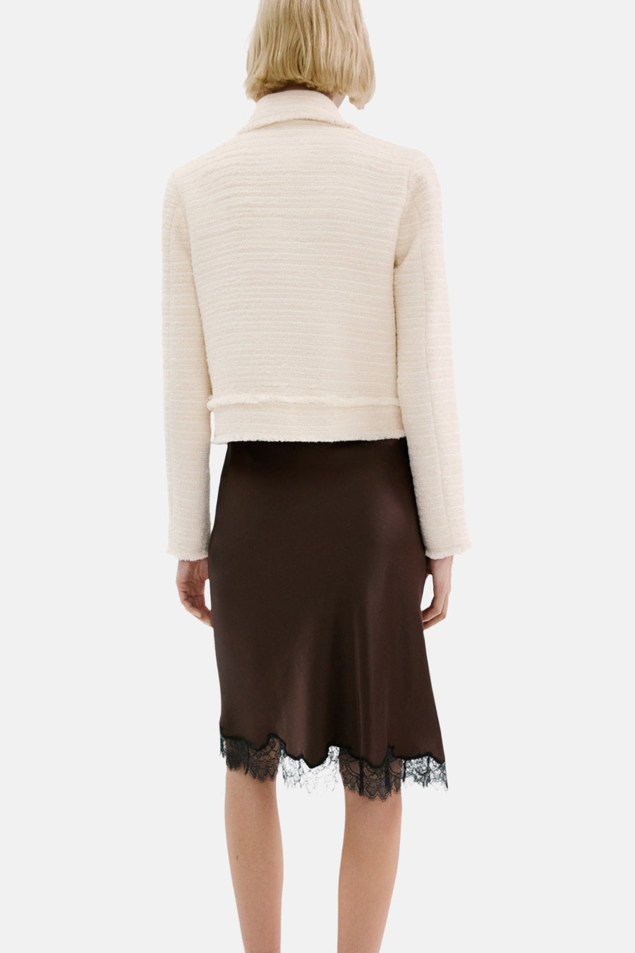 Een vrouw van achteren gezien, gekleed in een crèmekleurig jasje en een bruine rok met zwarte kanten rand.