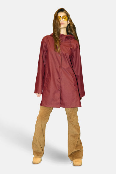 Bordeaux regenjas van DROPLETS, gedragen met een bruine corduroy broek en gele zonnebril.
