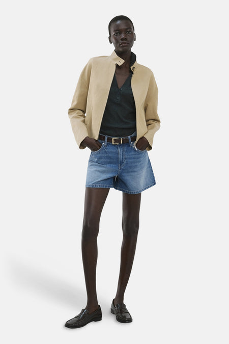 Middenblauw denim bermuda short van MANGO, gecombineerd met een beige jack en zwarte loafers.