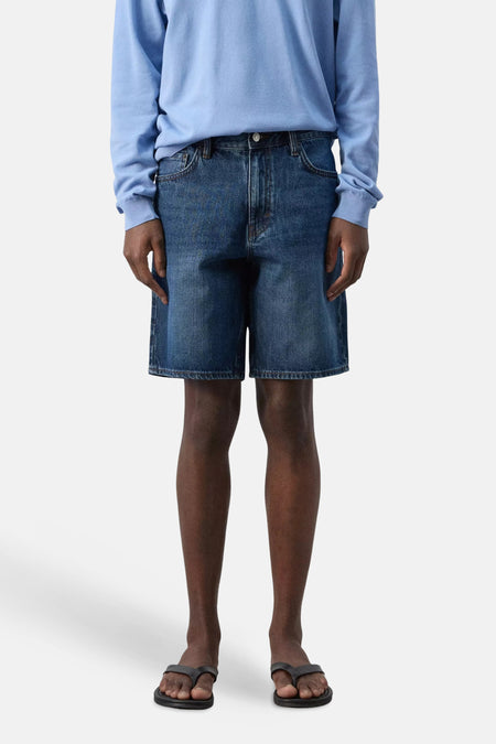 Donkerblauwe denim bermuda van MANGO, gedragen met een lichtblauw shirt en zwarte teenslippers.