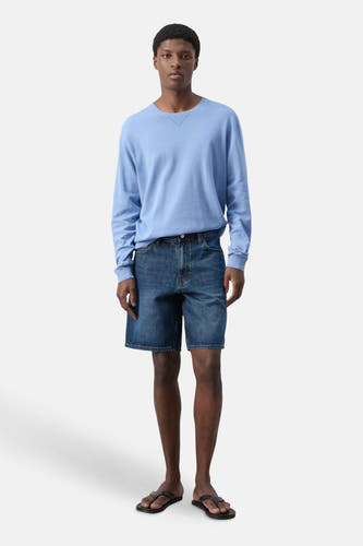 Donkerblauwe spijkerbermuda van MANGO, gedragen door een staande man in een lichtblauwe trui en zwarte sandalen.