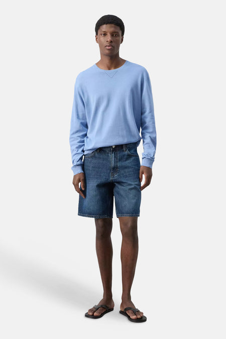 Donkerblauwe spijkerbermuda van MANGO, gedragen door een staande man in een lichtblauwe trui en zwarte sandalen.