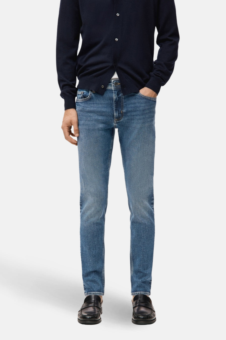 Slim fit jeans in middenblauw denim van MANGO, gedragen met een donkerblauw vest en zwarte loafers.