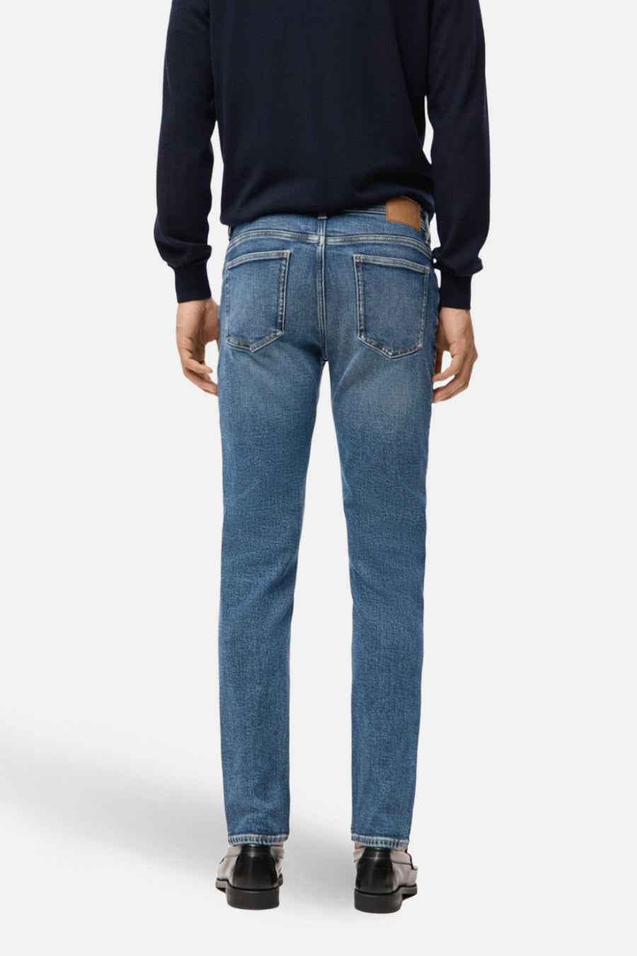Slim jeans in middenblauw denim van MANGO, gezien van achteren, gecombineerd met een donkerblauwe trui en zwarte schoenen.
