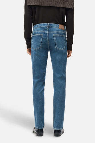 Taps toelopende jeans van donkerblauw denim van MANGO, achterkant gezien, met donker shirt en zwarte schoenen.
