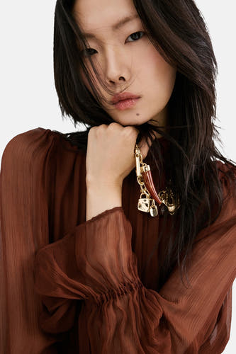 Robe marron à manches longues de MANGO, associée à un collier doré et un tissu transparent.