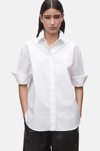 Chemise blanche à manches courtes de MANGO, avec manches retroussées et col classique, portée avec un pantalon noir.
