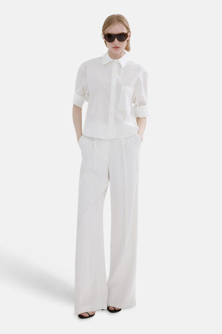Chemise blanche à manches longues de MANGO, portée avec un pantalon blanc ample et des tongs noires.