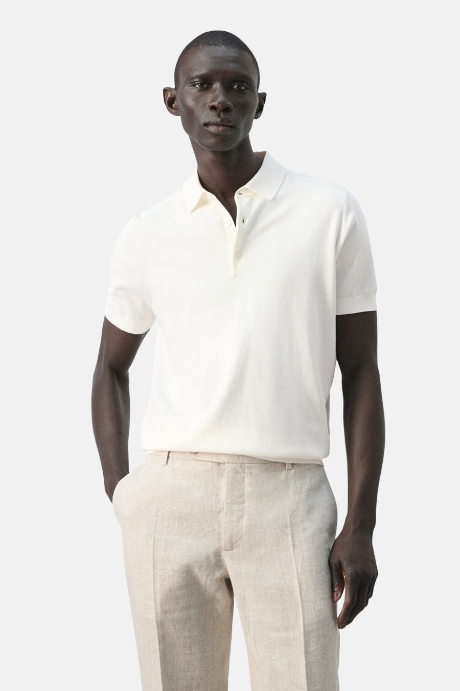 Witte polo van MANGO, gedragen met beige linnen broek, met korte mouwen en knoopsluiting.

