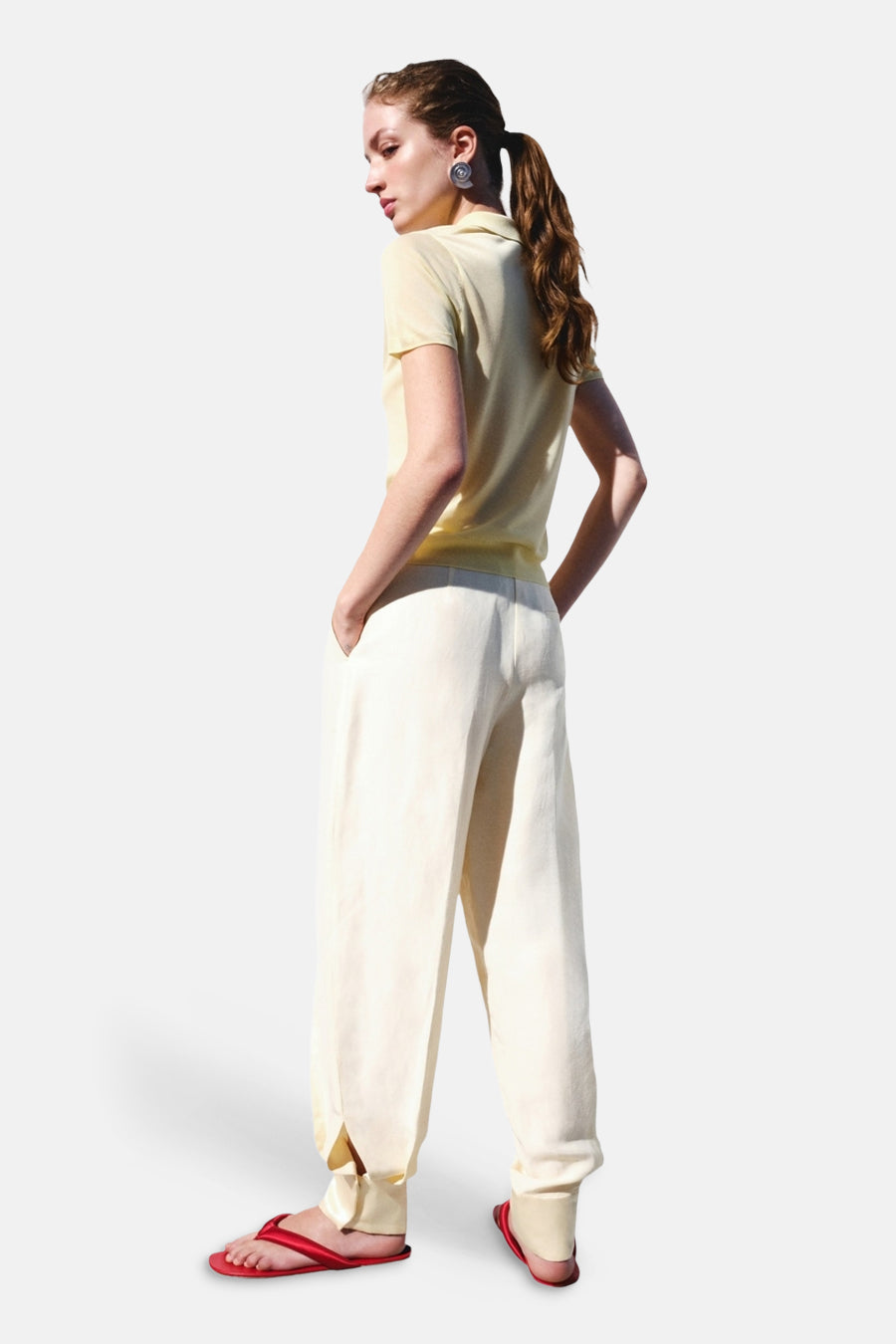 Gele polo met korte mouwen van MANGO, gedragen door een model met de rug naar de camera, gecombineerd met een off-white broek en rode slippers.