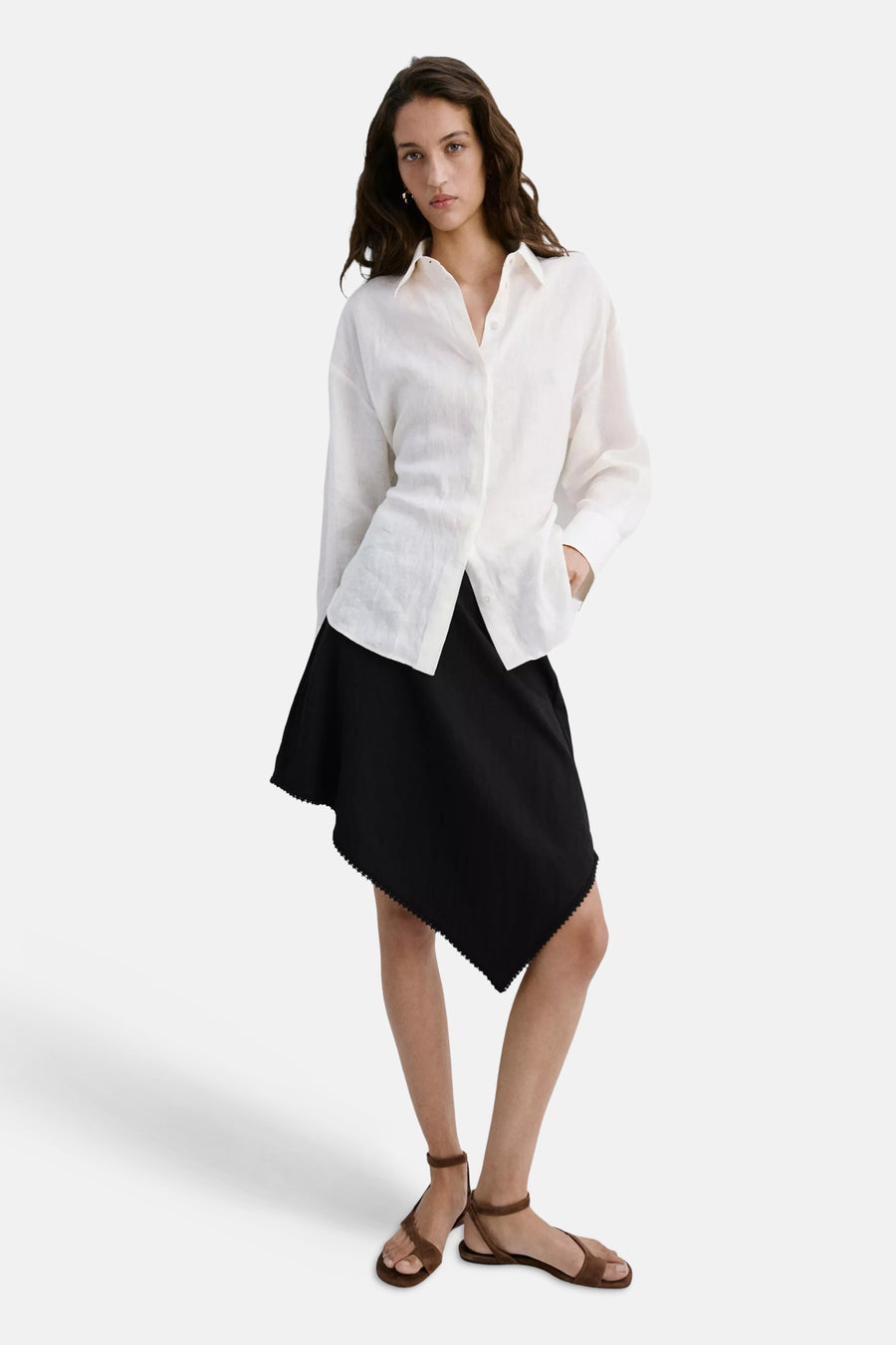 Zwarte rok van MANGO met asymmetrische zoom en decoratieve bies, gedragen met een wit overhemd en bruine sandalen.