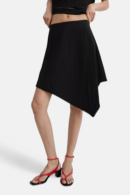 Zwarte rok van MANGO met een asymmetrische zoom en geschulpte rand, gedragen met een crop top en rode sandalen.