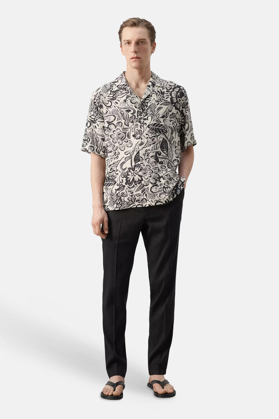 Chemise à manches courtes à motif floral noir et blanc de MANGO, portée par un mannequin avec un pantalon noir et des tongs.