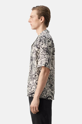 Chemise à manches courtes avec imprimé floral noir et blanc de MANGO, portée par un homme de profil avec un pantalon noir.