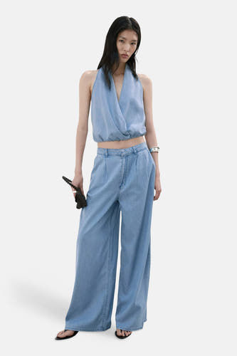 Lichtblauwe denim blouse zonder mouwen van MANGO, wrap-stijl met diep V-hals, gekenmerkt door een losse pasvorm en subtiele textuur, getoond in lifestyle-opstelling met bijpassende jeans en zwarte sandalen.