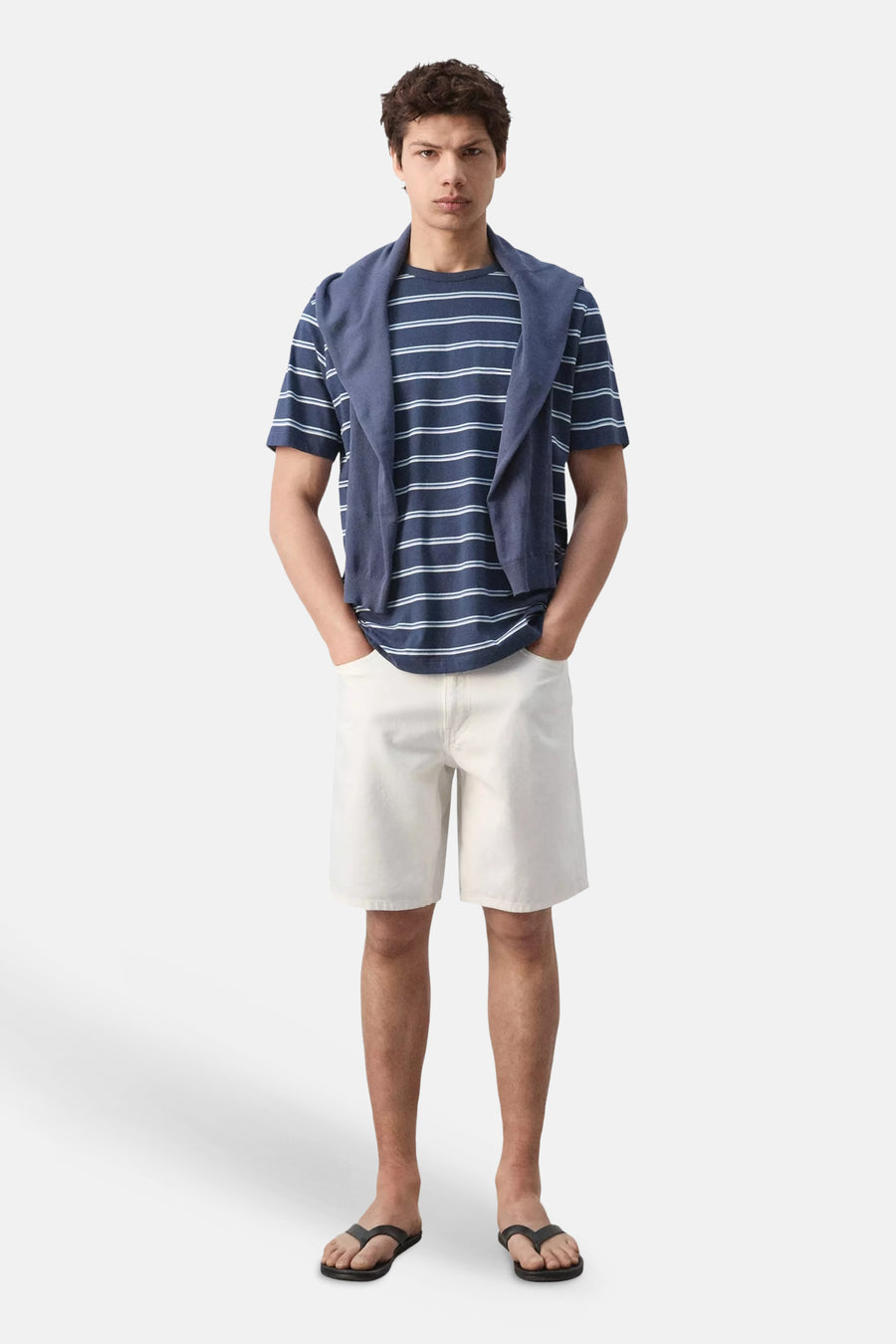T-shirt bleu rayé de MANGO, porté avec un pull sur les épaules, un short blanc et des tongs.
