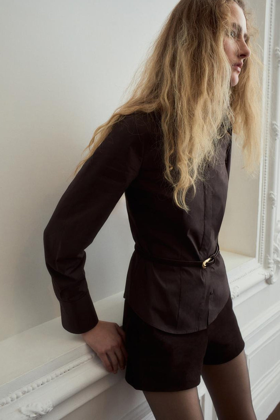 Chemise marron à manches longues de MANGO, cintrée avec une fine ceinture, combinée avec un short marron et des collants.