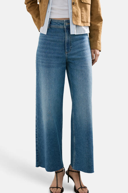 Wijde jeans van blauw denim van MANGO, met hoge taille en enkellange pijpen, gecombineerd met een bruine jas en sandalen.
