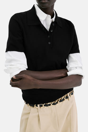 Polo noire à manches courtes de MANGO, portée avec une chemise blanche en dessous et une ceinture chaîne dorée.