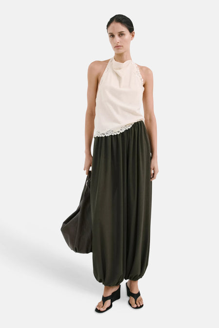 Haut sans manches écru à col licou, col drapé et bordure en dentelle de MANGO, portée avec un pantalon vert foncé et des sandales noires.