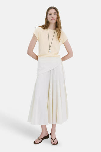 T-shirt jaune à manches courtes de MANGO, porté par un mannequin, stylisé avec une jupe portefeuille blanche et des sandales marron.