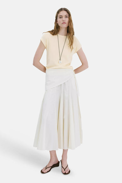 T-shirt jaune à manches courtes de MANGO, porté par un mannequin, stylisé avec une jupe portefeuille blanche et des sandales marron.