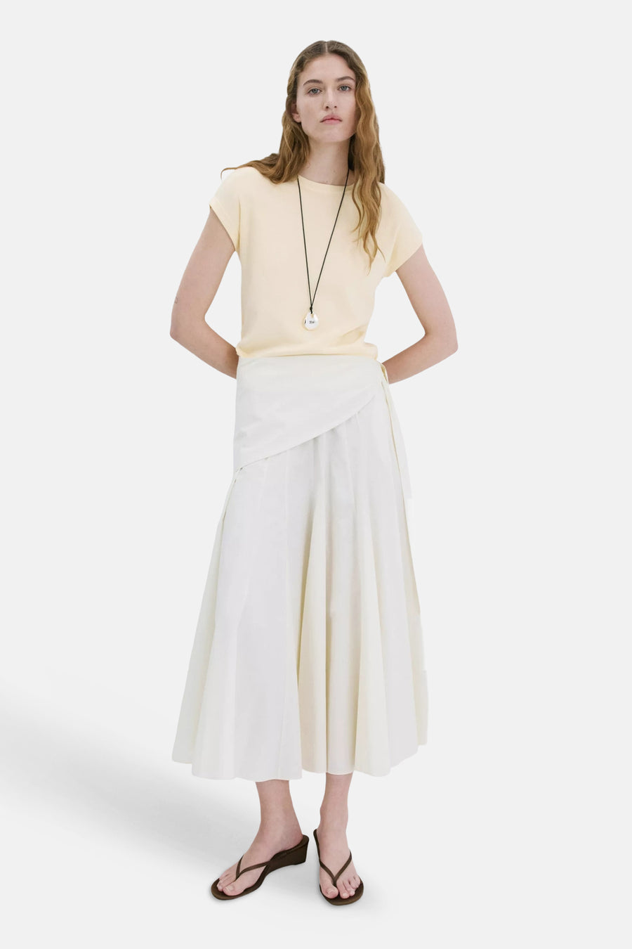 T-shirt jaune à manches courtes de MANGO, porté par un mannequin, stylisé avec une jupe portefeuille blanche et des sandales marron.