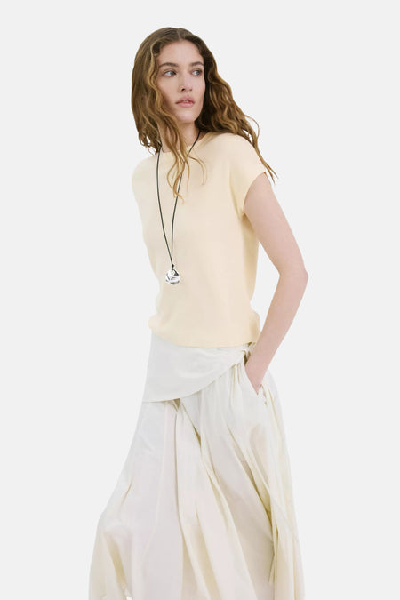 Haut jaune à manches courtes de MANGO, avec col rond, porté par un mannequin avec un pantalon blanc ample et un pendentif argenté.