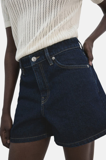 Donkerblauwe denim short van MANGO, met gele stiksels en een witte gebreide top.
