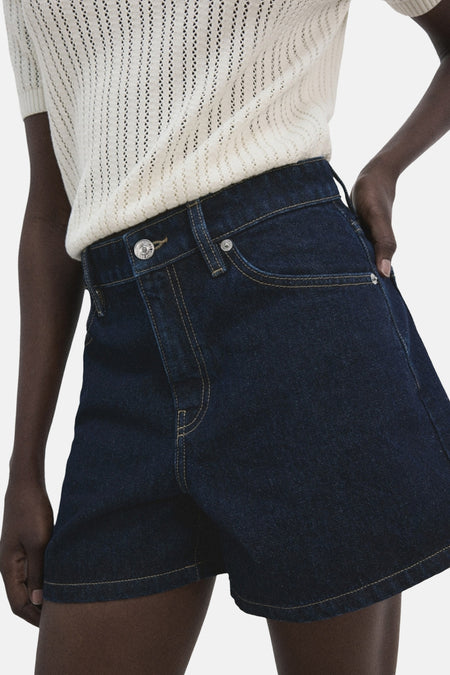 Donkerblauwe denim short van MANGO, met gele stiksels en een witte gebreide top.
