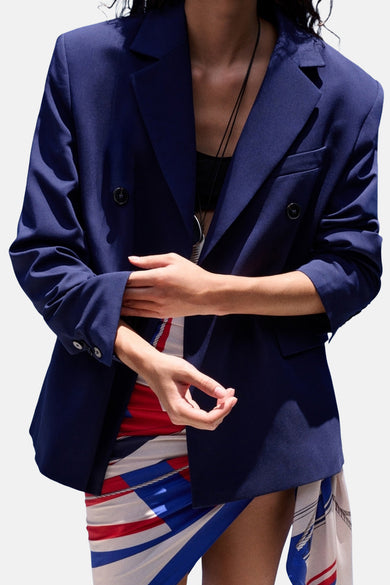Blazer bleu de MANGO, croisé et porté ouvert sur une jupe colorée avec manches retroussées.