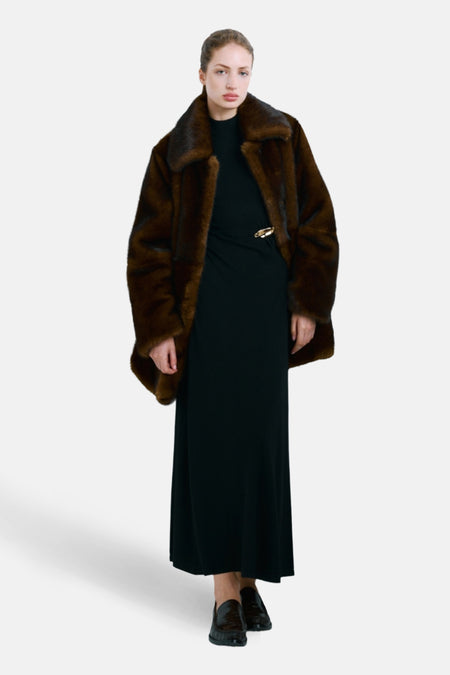 Vrouw in lange zwarte jurk en bruine faux fur jas, staand op een witte achtergrond.