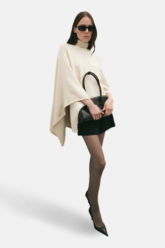 Vrouw met zonnebril, poncho, zwarte tas en zwarte schoenen.
