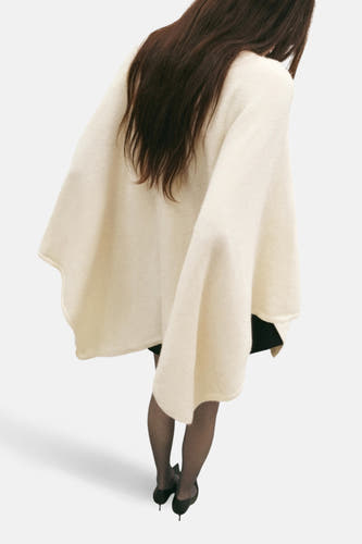 Ecru poncho van MANGO, met een losse pasvorm, gedragen over een zwarte jurk met panty en pumps.
