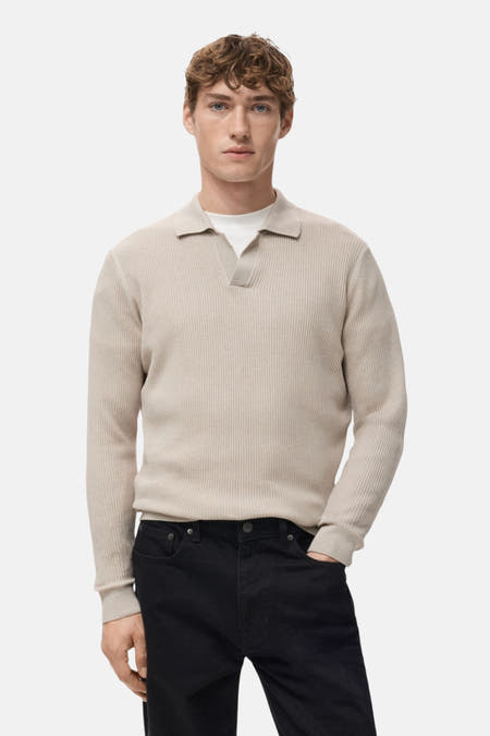 Beige V-hals trui van MANGO, met ribtextuur en gedragen over een wit T-shirt, met zwarte broek.
