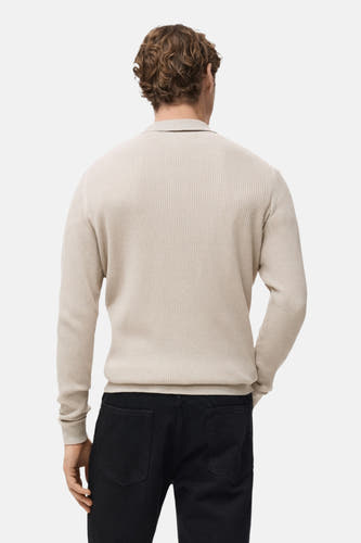 Beige V-hals trui van MANGO, met een ribgebreide textuur, gezien van de achterkant, gecombineerd met een zwarte broek.
