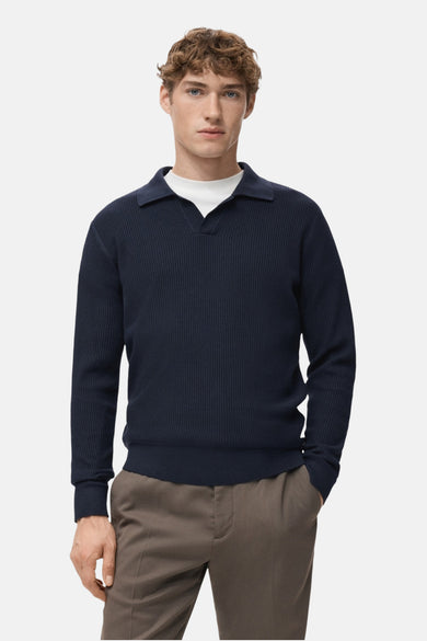 Pull à col V bleu de MANGO, avec une texture rayée et un col roulé sur un T-shirt blanc, associé à un pantalon marron.