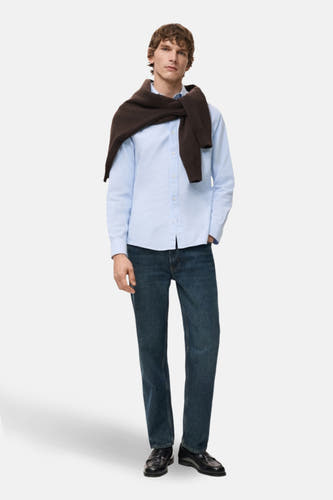 Chemise bleue à manches longues de MANGO, portée avec une veste marron sur les épaules.