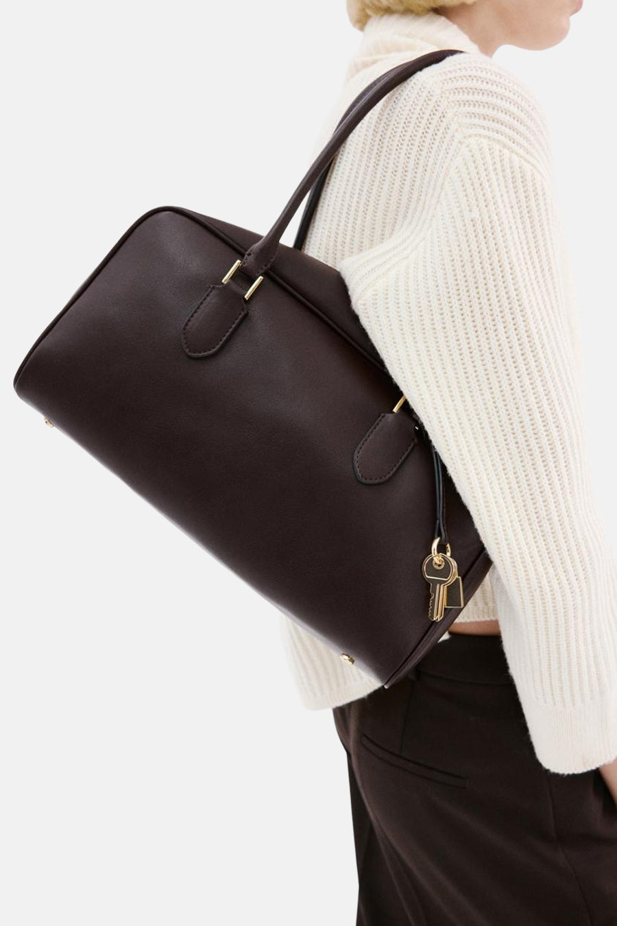 Bruine schoudertas van MANGO, met gouden details en een sleutelhanger, gedragen met een witte trui en bruine broek.
