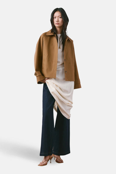 Veste courte marron de MANGO, portée sur une robe blanche et un jean foncé avec des talons.