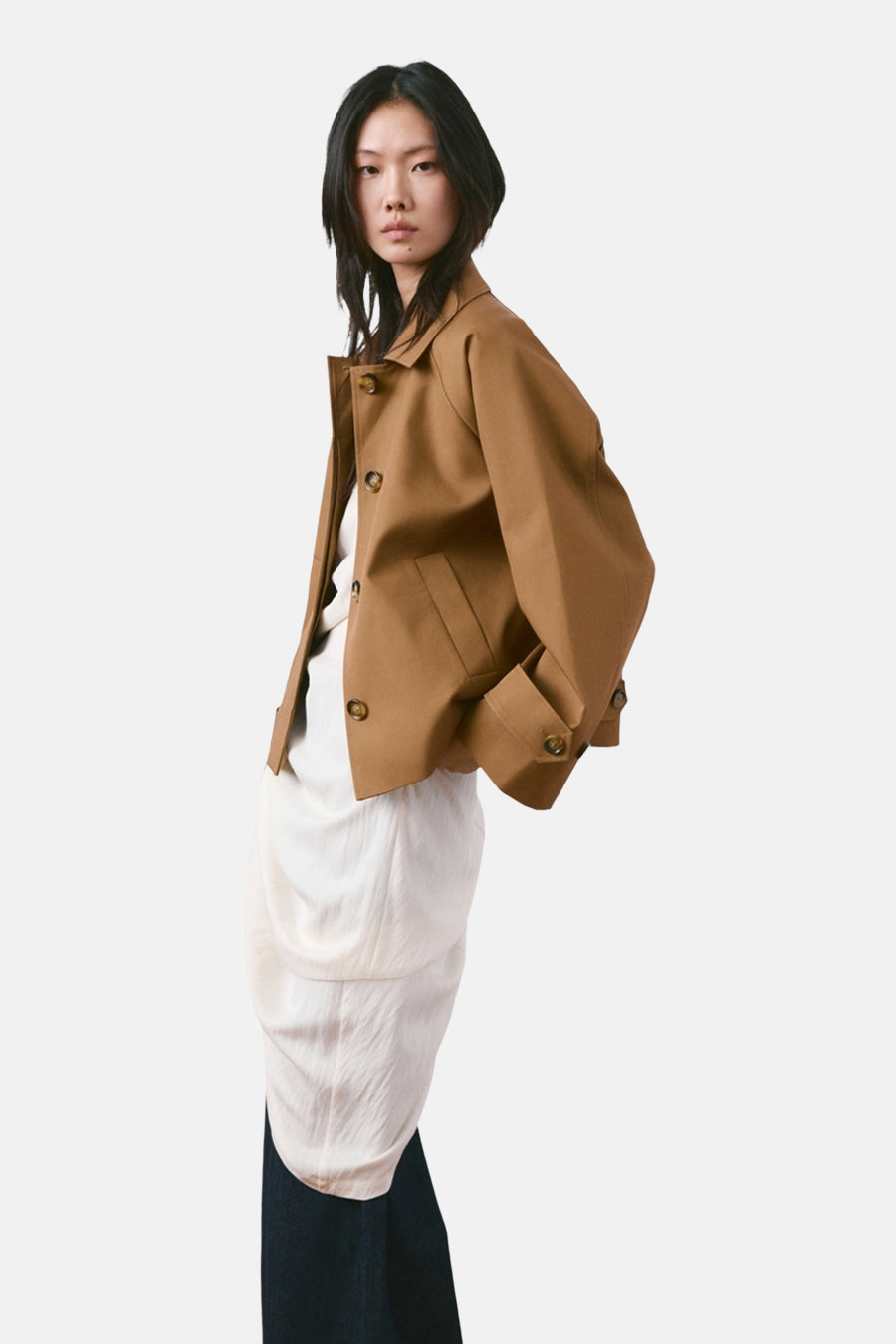 Veste courte marron de MANGO, avec fermeture à boutons, poches et manches longues ; portée sur une robe blanche et un pantalon foncé.