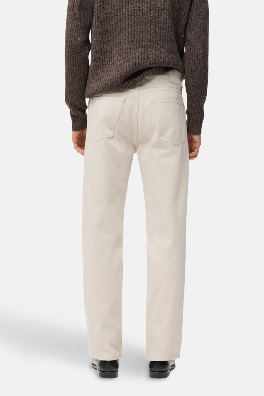Rechte jeans van off-white denim van MANGO, met achterzakken, gecombineerd met een bruine trui en zwarte schoenen, gezien van achteren.
