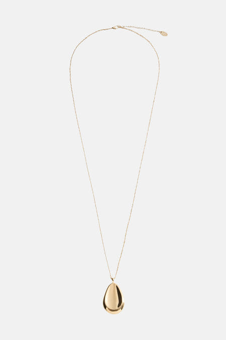 Long collier d'or de MANGO, avec un pendentif en forme de goutte, porté avec une chemise crème et une veste fausse fourrure marron.