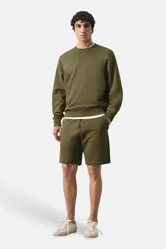 Pull kaki à col rond de MANGO, porté avec un t-shirt blanc en dessous et un short et des baskets assortis.