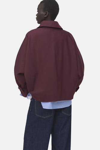 Korte bordeauxrode jas van MANGO, met kraag en lange mouwen, gedragen over een gestreept overhemd en donkerblauwe jeans.