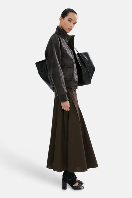 Jupe longue marron de MANGO, portée avec une veste en cuir et un cabas noir, sur un fond blanc.