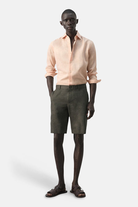 Bermuda de MANGO, combinée avec une chemise en lin rose clair et des sandales en cuir, debout sur un fond blanc.