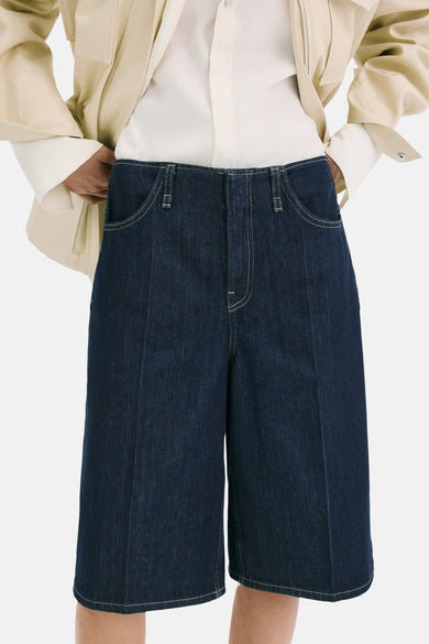 Bermuda en jean bleu marine de MANGO, vue de dos, combinée avec une veste de couleur crème et des sandales noires.