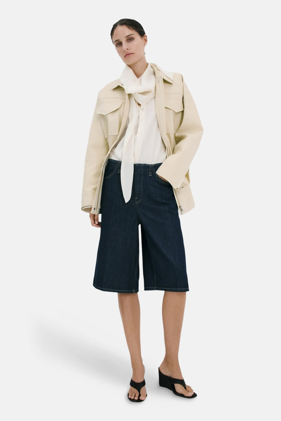 Bermuda en denim bleu marine de MANGO, combinée avec une veste crème et une blouse blanche à nœud.